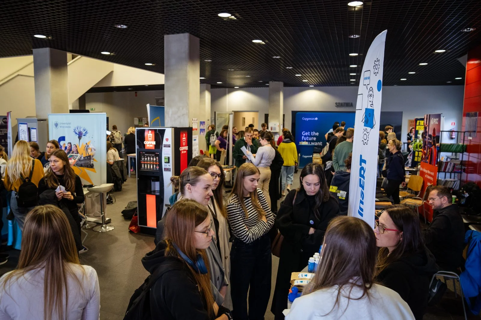 Zdjęcie wykonane wewnątrz dużej, nowoczesnej hali. Widoczna jest duża liczba osób stojących i rozmawiających przy stoiskach wystawienniczych. 