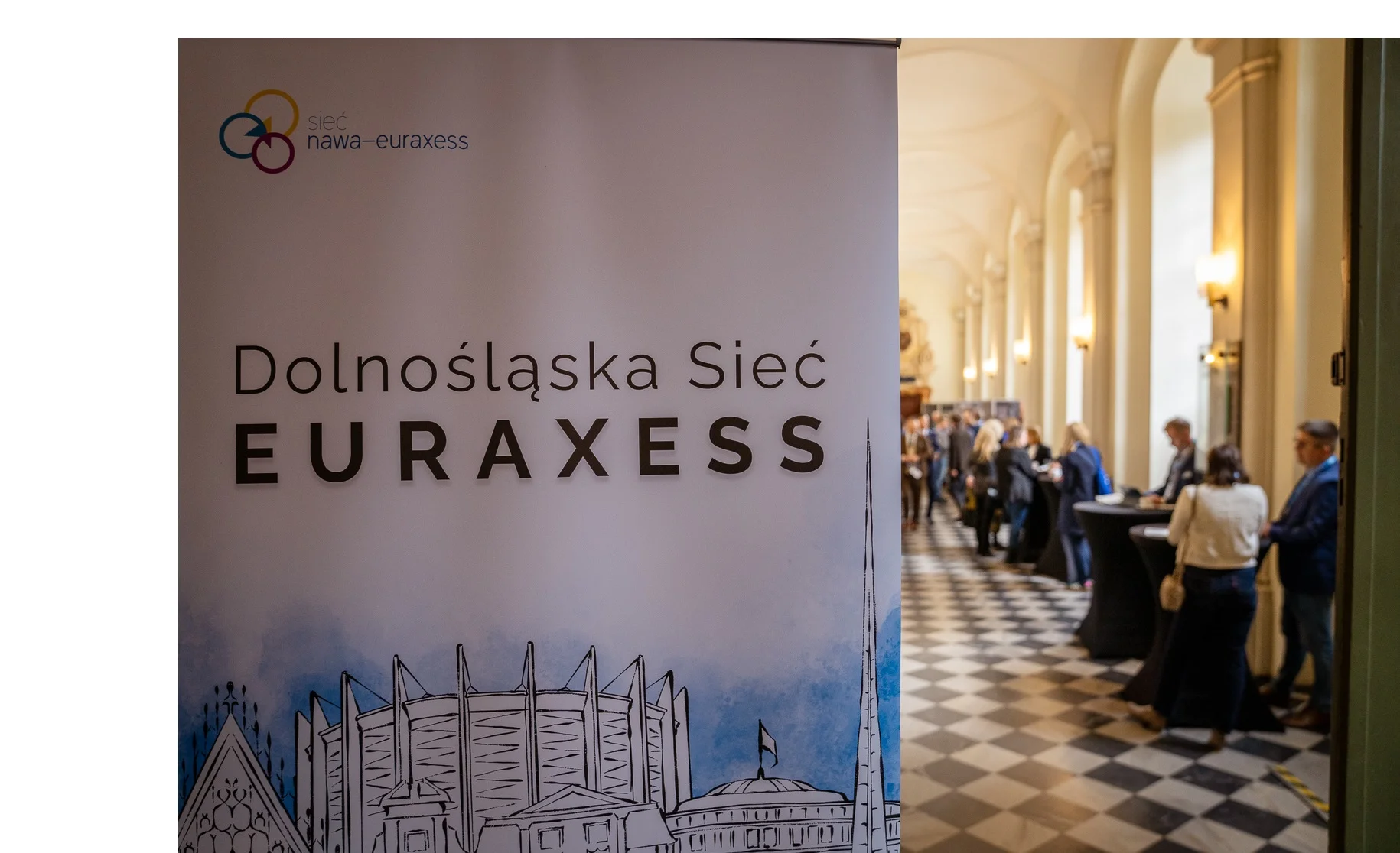 Zdjęcie wykonane we wnętrzu wysokiego, reprezentacyjnego korytarza z jasnymi kolumnami i sklepionym sufitem i podłogą z wzorem szachownicy.
Na pierwszym planie po lewej stronie widoczny jest wysoki pionowy baner z napisem Dolnośląska Sieć EURAXESS oraz logotypem sieć nawa euraxess. Dolna część baneru zawiera stylizowaną graficzną ilustrację budynków ratusza miejskiego, hali stulecia i panoramy racławickiej.
W tle, po prawej stronie, znajduje się grupa osób stojących przy wysokich stolikach, ustawionych wzdłuż korytarza. Wzdłuż ścian widoczne są kolumny oraz punktowe oświetlenie. 