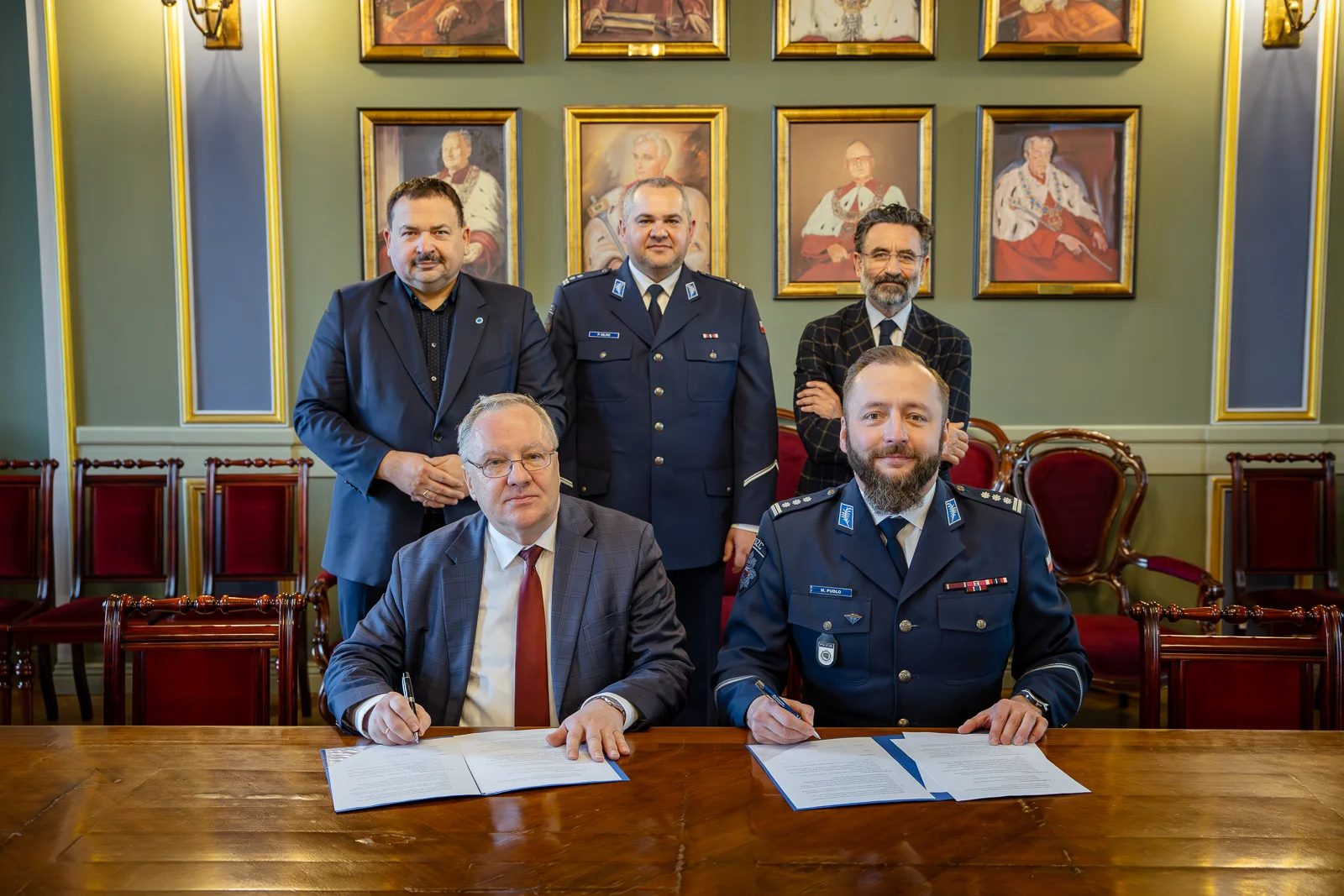 Zdjęcie wykonane w reprezentacyjnej sali z zielonymi ścianami, złotymi ornamentami i portretami w ozdobnych ramach zawieszonymi w tle. W pomieszczeniu znajduje się długi, drewniany stół.
Na pierwszym planie, przy stole, siedzą dwie osoby podpisujące dokumenty rozłożone na blatach. Obie osoby trzymają długopisy, a przed każdą z nich leży otwarty dokument. Jedna osoba ubrana jest w garnitur, druga w mundur służbowy.
Za siedzącymi osobami stoją cztery inne osoby ustawione w jednym rzędzie. Dwie z nich mają na sobie mundury, pozostałe noszą stroje formalne.
W tle widoczne są czerwone, tapicerowane krzesła ustawione wzdłuż ściany. Nad nimi wiszą liczne portrety postaci historycznych w złotych ramach. Oświetlenie jest ciepłe i równomierne, podkreślające oficjalny charakter wnętrza. Fotografia dokumentuje formalne wydarzenie o charakterze urzędowym lub ceremonialnym.