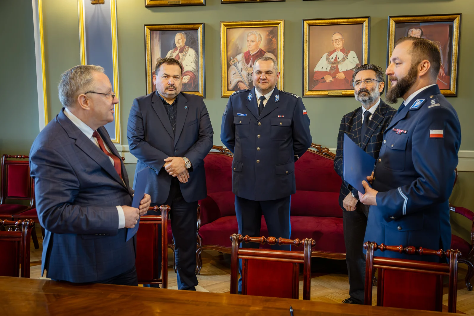 Zdjęcie wykonane w reprezentacyjnej sali z zielonymi ścianami, złotymi zdobieniami i portretami w ozdobnych ramach zawieszonymi w tle. W pomieszczeniu znajduje się długi drewniany stół oraz czerwone, tapicerowane krzesła ustawione wzdłuż ściany.
Na pierwszym planie, po lewej stronie kadru, stoi osoba w ciemnym garniturze, zwrócona w stronę grupy pozostałych osób. Po prawej stronie znajduje się osoba w mundurze służbowym trzymająca teczkę z dokumentami.
Pomiędzy nimi, w głębi kadru, stoją cztery inne osoby ubrane w stroje formalne i mundury, ustawione w półokręgu przy ścianie.
W tle widoczna jest sofa obita czerwonym materiałem, a nad nią kilka portretów postaci historycznych w złotych ramach. Oświetlenie jest równomierne i ciepłe, podkreślające oficjalny i ceremonialny charakter wnętrza. Fotografia dokumentuje spotkanie o charakterze formalnym w pomieszczeniu reprezentacyjnym.