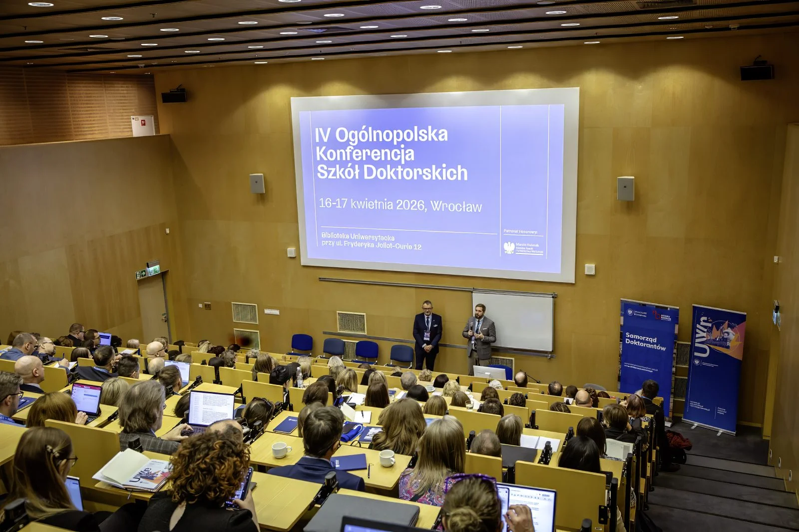 Zdjęcie wykonane w dużej auli wykładowej z drewnianymi ścianami i rzędami jasnożółtych, amfiteatralnie ustawionych ławek. Sala jest wypełniona uczestnikami siedzącymi przy stolikach, na których znajdują się laptopy, notesy oraz materiały konferencyjne.
Na froncie sali, przy dolnej krawędzi ekranu projekcyjnego, stoją dwie osoby ubrane formalnie. Nad nimi znajduje się duży ekran z prezentacją. Na slajdzie widnieje tekst: IV Ogólnopolska Konferencja Szkół Doktorskich, 16–17 kwietnia 2026, Wrocław. W dolnej części slajdu widoczne są logotypy organizatorów.
Po prawej stronie sali stoją pionowe roll‑upy informacyjne, w tym jeden z napisem Samorząd Doktorantów oraz drugi z logotypem Uniwersytetu Wrocławskiego.
Uczestnicy wypełniają większość miejsc w sali i są skierowani w stronę ekranu oraz osób stojących z przodu. Oświetlenie jest jasne, równomierne, a zdjęcie dokumentuje przebieg wydarzenia konferencyjnego w przestrzeni akademickiej.