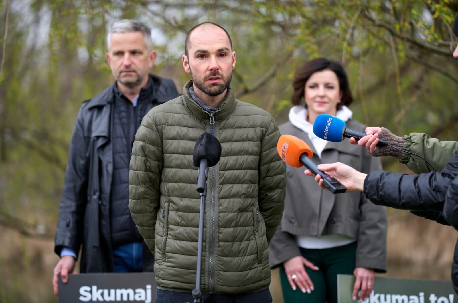 Zdjęcie wykonane w plenerze, w zielonym otoczeniu z drzewami i trawą. Na pierwszym planie pośrodku kadru stoi osoba ubrana w pikowaną, oliwkową kurtkę, przed mikrofonem umieszczonym na statywie.
Z prawej strony kadru w kierunku osoby skierowane są ręce trzymające mikrofony reporterskie w kolorach pomarańczowym i niebieskim.
W tle widoczne są trzy inne osoby stojące w pewnym oddaleniu. Dwie z nich trzymają pionowe plansze informacyjne z napisem Skumaj to! oraz zdjęciami płazów. Na jednej planszy można odczytać fragment tekstu: Żaba moczarowa na wrocławskich mokradłach.
Tło zdjęcia stanowi naturalny krajobraz z drzewami i krzewami.
