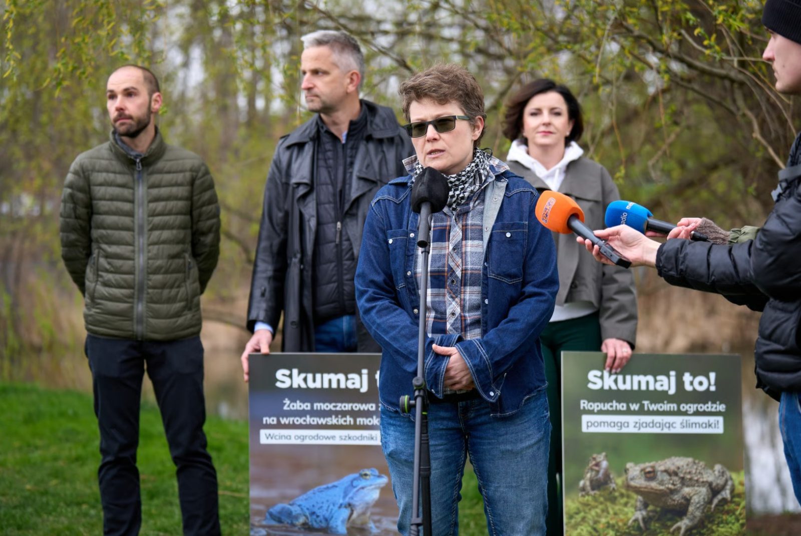 Zdjęcie wykonane na zewnątrz, w zielonej przestrzeni z drzewami i trawą, wczesną wiosną. Na pierwszym planie, pośrodku kadru, znajduje się osoba stojąca przed mikrofonem umieszczonym na statywie. Osoba ubrana jest w dżinsową kurtkę, koszulę w kratę i dżinsowe spodnie oraz nosi ciemne okulary.
Z prawej strony kadru w stronę osoby skierowane są ręce trzymające mikrofony reporterskie w kolorach pomarańczowym i niebieskim.
W tle, za osobą stojącą przy mikrofonie, znajduje się kilka innych osób ustawionych w półokręgu. Każda z nich trzyma plansze informacyjne. Na jednej z plansz widoczny jest napis: Skumaj to! Żaba moczarowa na wrocławskich mokradłach wraz ze zdjęciem żaby. Na drugiej planszy znajduje się tekst: Skumaj to! Ropucha w Twoim ogrodzie pomaga zjadać ślimaki! oraz zdjęcie ropuchy.
Tło fotografii stanowią drzewa z młodymi liśćmi oraz naturalny krajobraz, bez zabudowy.