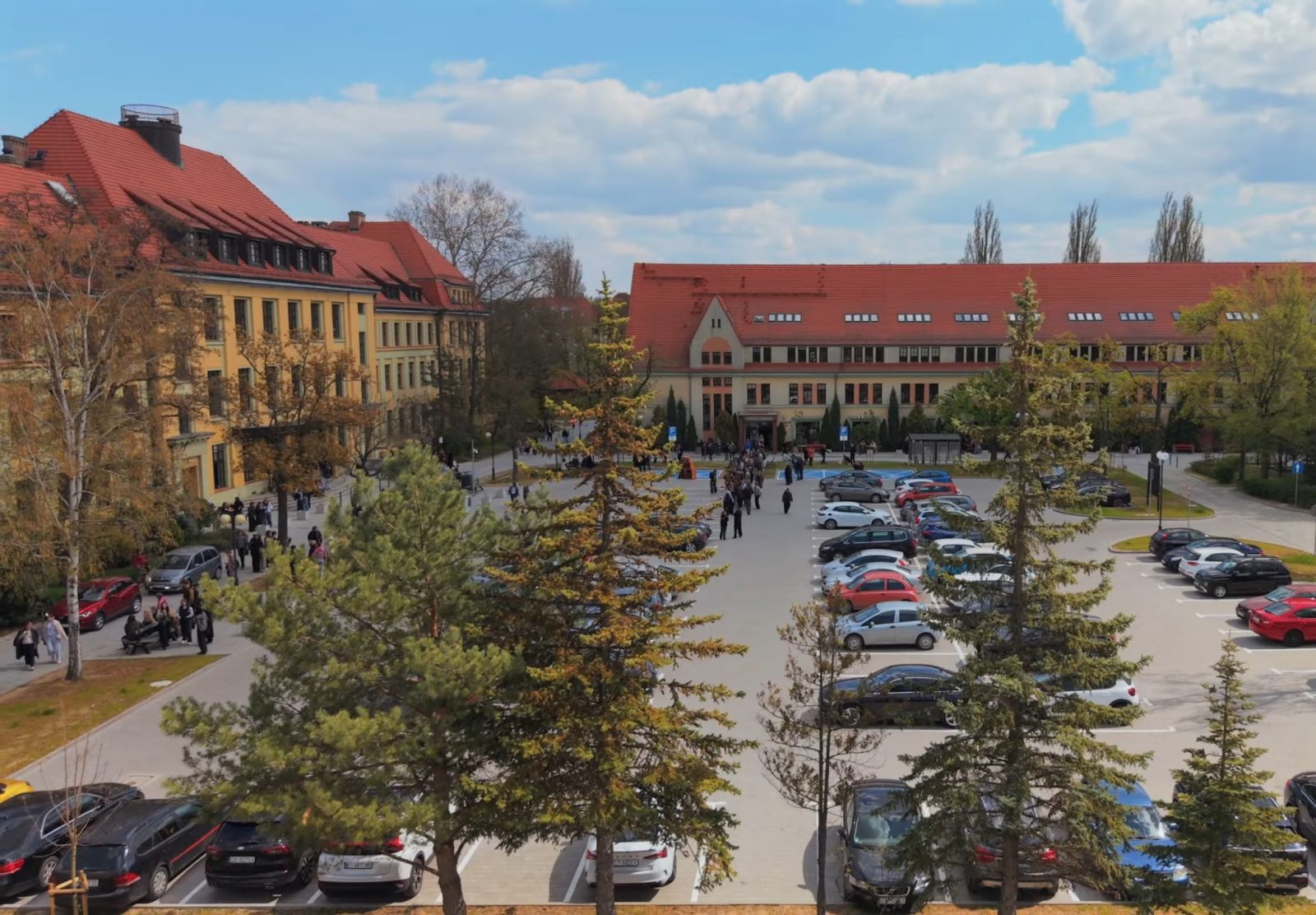 Panoramiczne zdjęcie kampusu Uniwersytetu Wrocławskiego wykonane z podwyższonej perspektywy. Na pierwszym planie widoczny jest duży parking z zaparkowanymi samochodami oraz alejki piesze obsadzone drzewami i krzewami. Między alejkami poruszają się grupy pieszych.
W tle znajdują się zabudowania uczelni z charakterystycznymi jasnymi elewacjami i czerwonymi dachami, w tym długi budynek centralny z wejściem głównym. Po lewej stronie widoczny jest kolejny gmach o podobnej architekturze. Nad kampusem rozciąga się jasne, lekko zachmurzone niebo.