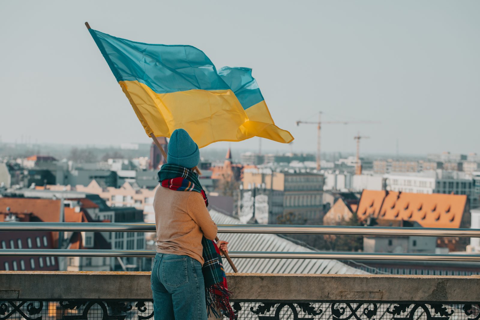 osoba trzymająca flagę ukrainy na drzewcu na balkonie z widokiem na panoramę miasta