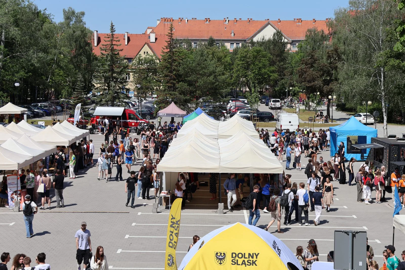 Zdjęcie wykonane z podwyższonej perspektywy przedstawia duży, otwarty plac wypełniony stoiskami i uczestnikami wydarzenia plenerowego. Na środku ustawiono rzędy jasnych namiotów targowych, pod którymi znajdują się stanowiska informacyjne i stoiska.
Między namiotami porusza się duża liczba osób, część stoi w grupach przy stoiskach, inni przemieszczają się alejkami. Po prawej stronie widoczny jest niebieski namiot oraz food truck, przy którym stoją ludzie.
W dolnej części kadru znajduje się żółty namiot z napisem Dolny Śląsk. W centralnej części placu ustawiony jest czerwony pojazd służb oraz kilka mniejszych stoisk i banerów.
W tle widoczny jest parking z licznymi samochodami oraz otaczająca go zieleń – drzewa i krzewy. Za parkingiem stoją budynki o jasnych elewacjach i czerwonych dachach.