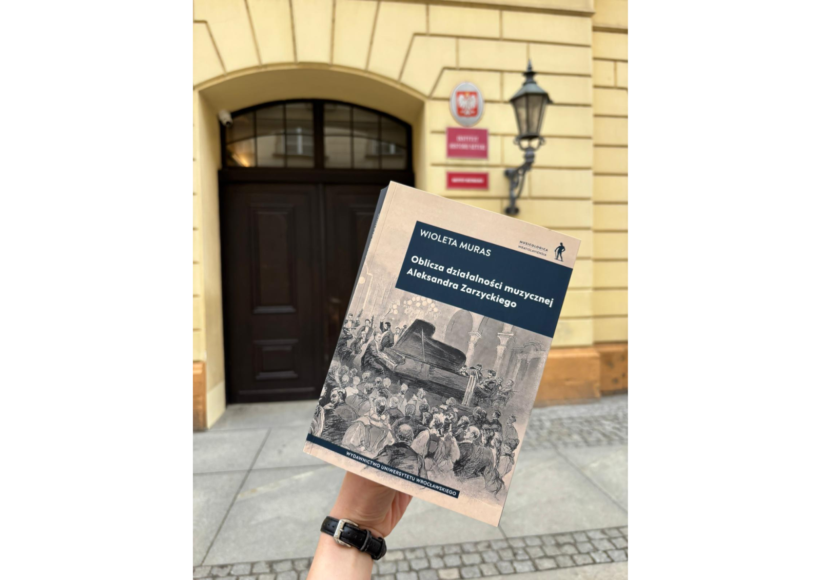 Zdjęcie przedstawia fragment elewacji jasnego, historycznego budynku z łukowym wejściem i ciemnymi drewnianymi drzwiami. Nad drzwiami znajduje się godło Polski oraz tabliczka z napisem Instytut Muzykologii. Obok drzwi zamocowana jest klasyczna latarnia ścienna.
Na pierwszym planie widoczna jest dłoń trzymająca książkę. Na okładce książki znajduje się napis: Wioleta Muras – Oblicza działalności muzycznej Aleksandra Zarzyckiego. Dolną część okładki zdobi czarno‑biała ilustracja przedstawiająca scenę koncertową z fortepianem i publicznością. U dołu widnieje napis: Wydawnictwo Uniwersytetu Wrocławskiego.