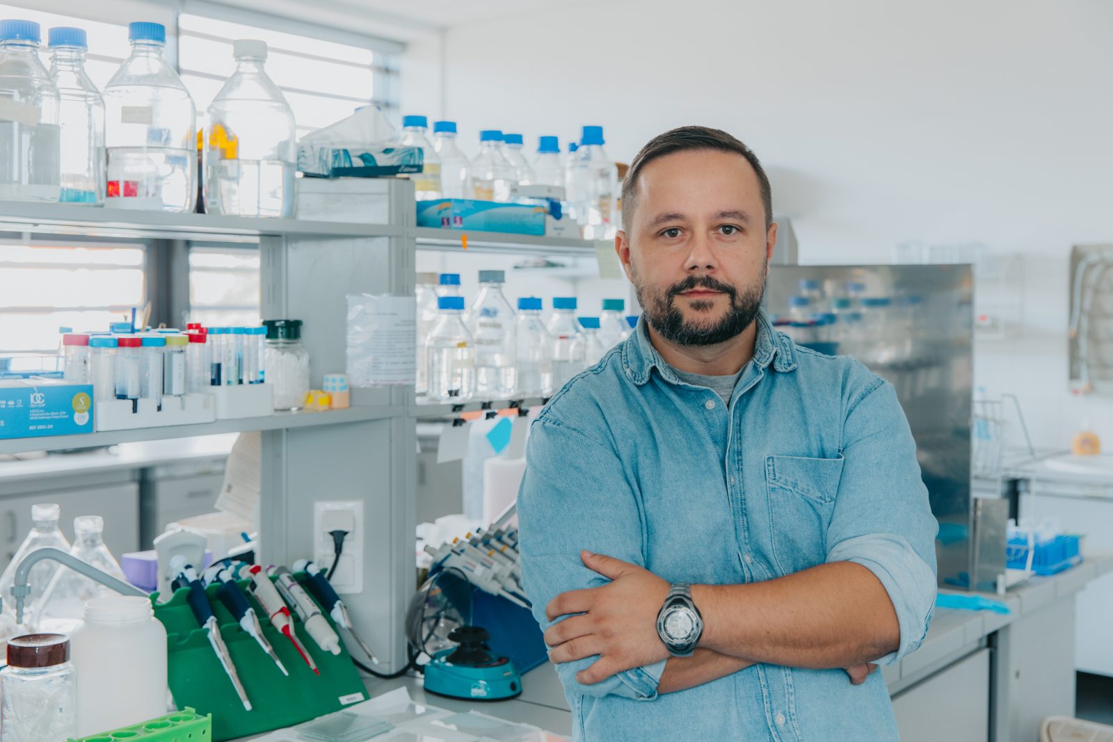 Zdjęcie wykonane we wnętrzu laboratorium. Na pierwszym planie, po prawej stronie kadru, stoi osoba ubrana w jasnoniebieską koszulę z podwiniętymi rękawami, z rękami skrzyżowanymi na wysokości klatki piersiowej. Na lewym nadgarstku widoczny jest zegarek.
W tle znajduje się stanowisko laboratoryjne z półkami i blatem roboczym. Na półkach ustawione są liczne przezroczyste butelki i pojemniki z cieczami, w większości zamknięte niebieskimi nakrętkami. Obok znajdują się probówki w stojakach oraz pudełka z materiałami laboratoryjnymi.
Na blacie widoczne są pipety automatyczne ułożone w stojaku, wirówka laboratoryjna, pojemniki i akcesoria do pracy badawczej.
