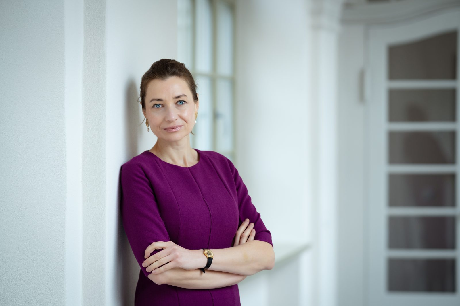 Na zdjęciu widoczna jest kobieta stojąca w przestronnym, dobrze oświetlonym wnętrzu. Ma ciemne włosy upięte do tyłu i ubrana jest w jednolitą sukienkę w odcieniu fioletu. Stoi lekko oparta o ścianę, z rękami skrzyżowanymi na wysokości talii. Na nadgarstku ma zegarek, a w uszach niewielkie kolczyki.