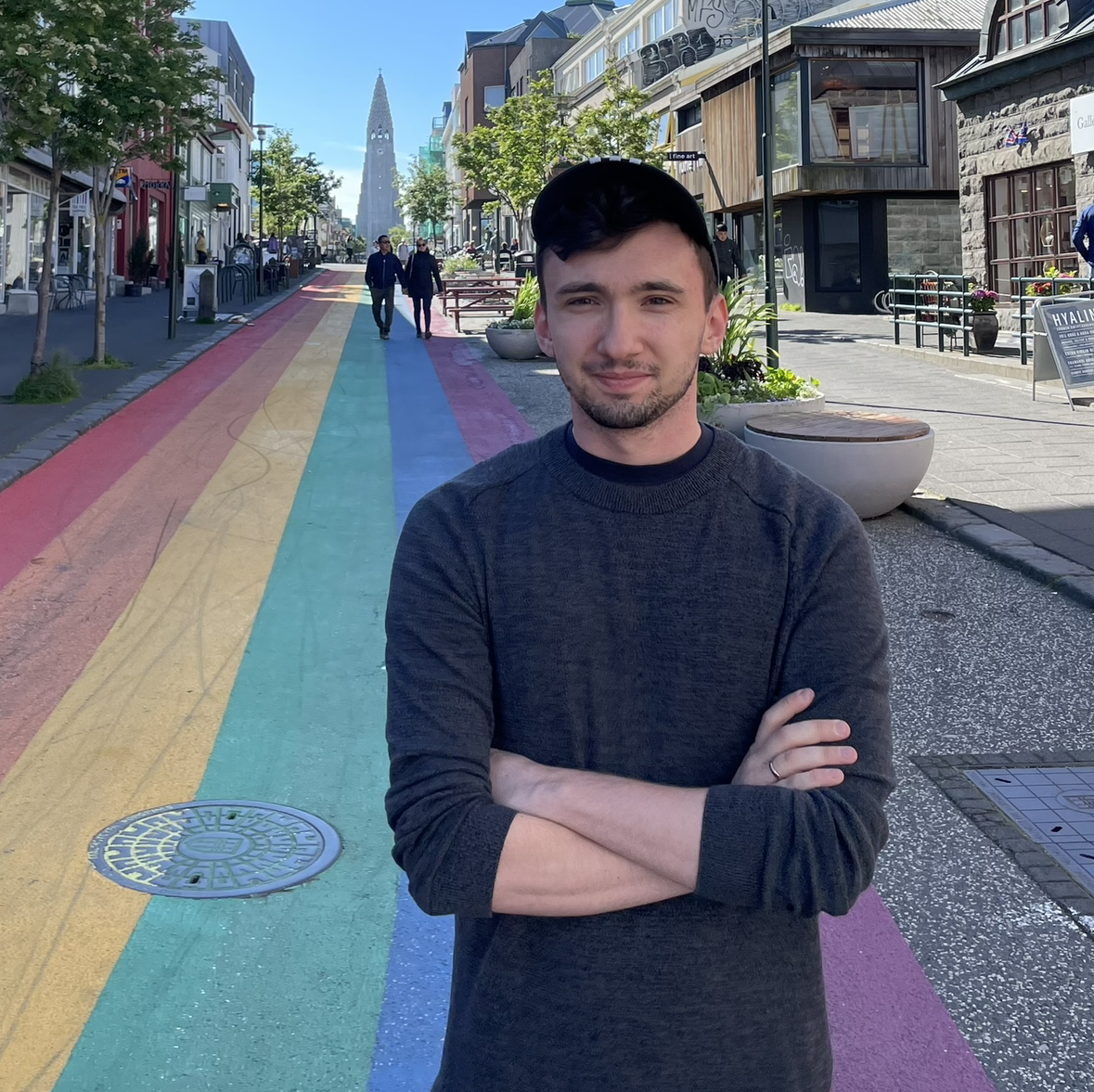 Zdjęcie przedstawia osobę stojącą na szerokiej ulicy przeznaczonej dla pieszych. Nawierzchnia ulicy pomalowana jest w pionowe pasy w kolorach tęczy: czerwonym, pomarańczowym, żółtym, zielonym, niebieskim i fioletowym.
Na pierwszym planie, centralnie, znajduje się osoba ubrana w ciemny sweter i czapkę z daszkiem, stojąca z rękami skrzyżowanymi.
W tle widoczna jest miejska zabudowa z budynkami po obu stronach ulicy. Po prawej stronie znajdują się donice z roślinami oraz ławki, po lewej sklepy i witryny. Na ulicy znajdują się także piesi przechodzący w oddali.