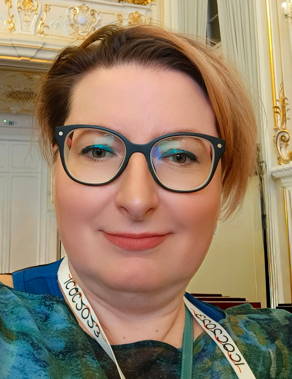 Zbliżenie twarzy osoby w okularach. W tle widoczne jest ozdobne wnętrze z jasnymi ścianami, dekoracyjnymi złotymi elementami oraz zasłonami. Na pierwszym planie widoczna jest twarz wypełniająca większość kadru, a przy szyi znajduje się pasek lub smycz z nadrukiem