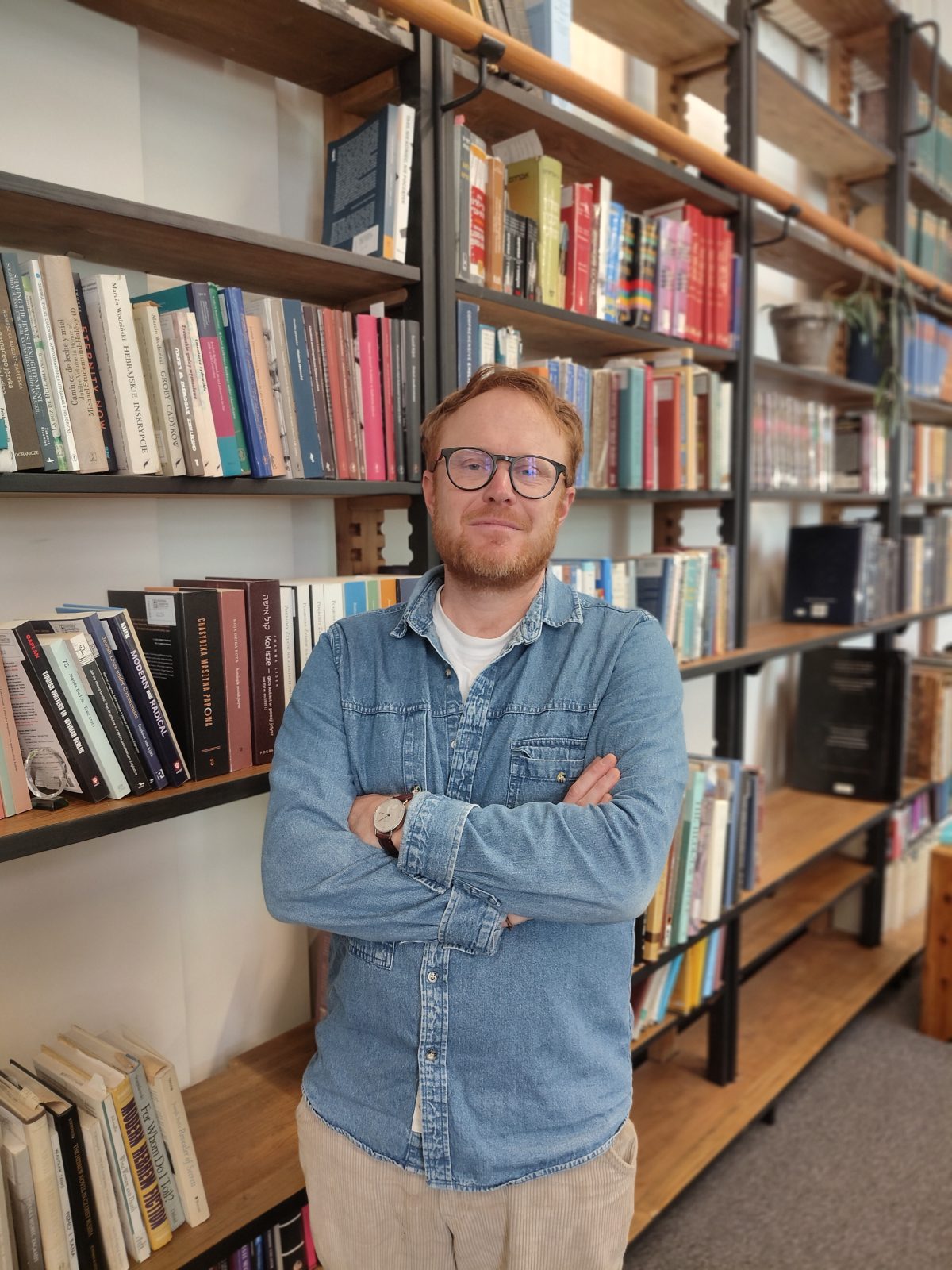 Na zdjęciu widoczna jest osoba stojąca na tle dużych regałów bibliotecznych sięgających od podłogi niemal do sufitu. Regały wykonane są z drewna i metalu oraz wypełnione książkami o różnorodnych kolorach i formatach. Mężczyzna ma krótkie rude włosy, okulary oraz rudawy zarost. Ubrany jest w jasnoniebieską koszulę dżinsową narzuconą na biały T-shirt. Stoi z rękami skrzyżowanymi na piersi, zwrócony przodem do aparatu