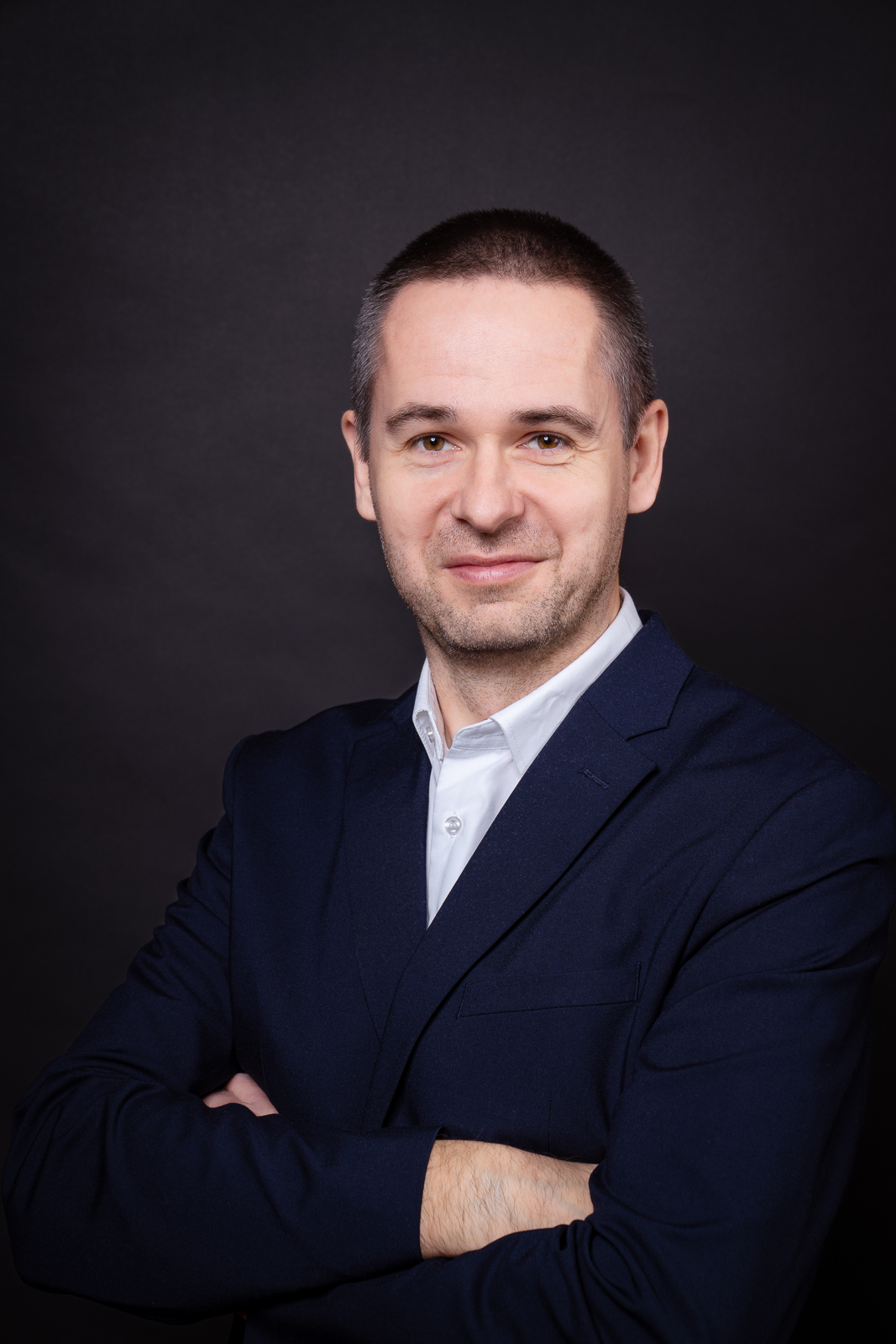 mężczyzna w garniturze i białej koszuli stojący w ciemnym pomieszczeniu z założonymi rękami